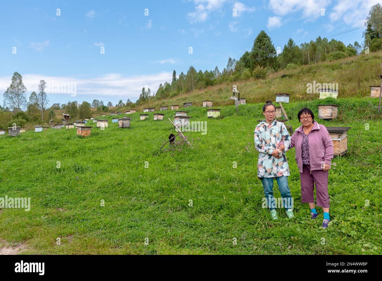 An Asian Yakut family an elderly mother and a young daughter pose hand in hand against the background of hives for bees in an apiary in the Altai moun Stock Photo