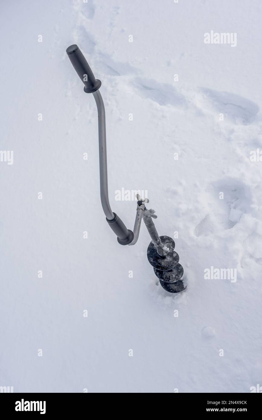 An iron black hand-held ice drill with a curved handle and screws frozen in ice stands screwed into the ice in winter against a background of white sn Stock Photo