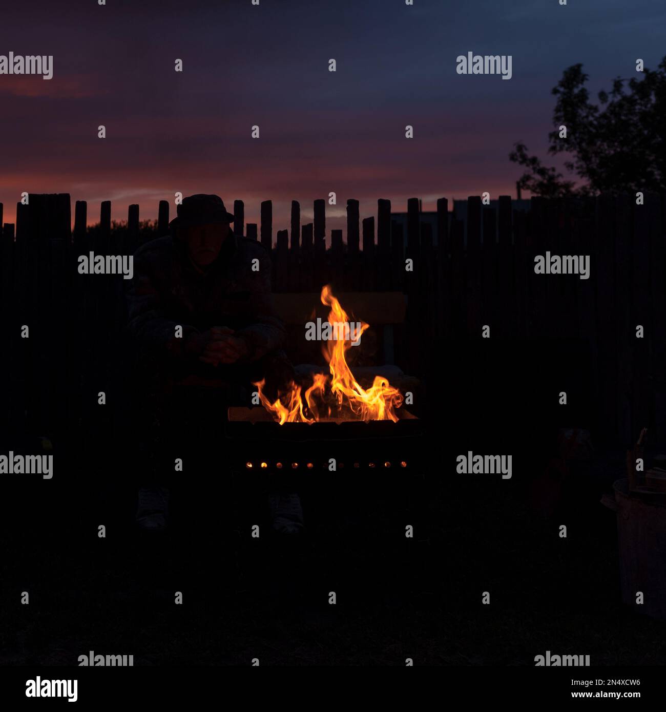 A man in the shade looks at the flames of the fire in the grill at night against the background of the setting sun at sunset in the village behind the Stock Photo