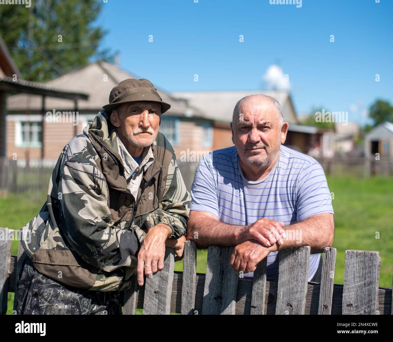 Two Russian village neighbors stand posing in conversation at the fence against the background of houses in the summer. Stock Photo