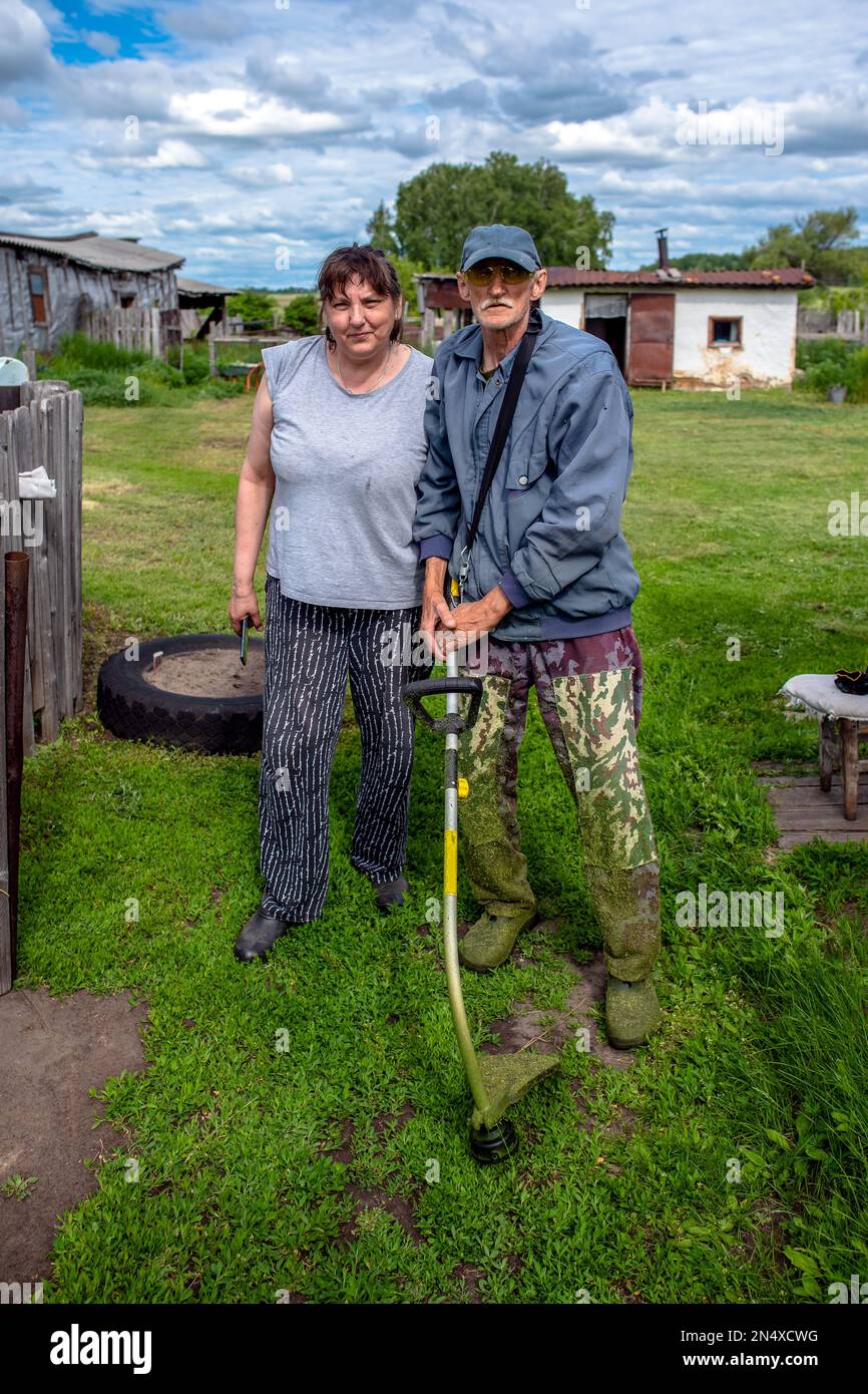 Family in the garden in the village of a stout woman and a thin brother with a grass trimmer stand on the lawn in the summer. Stock Photo