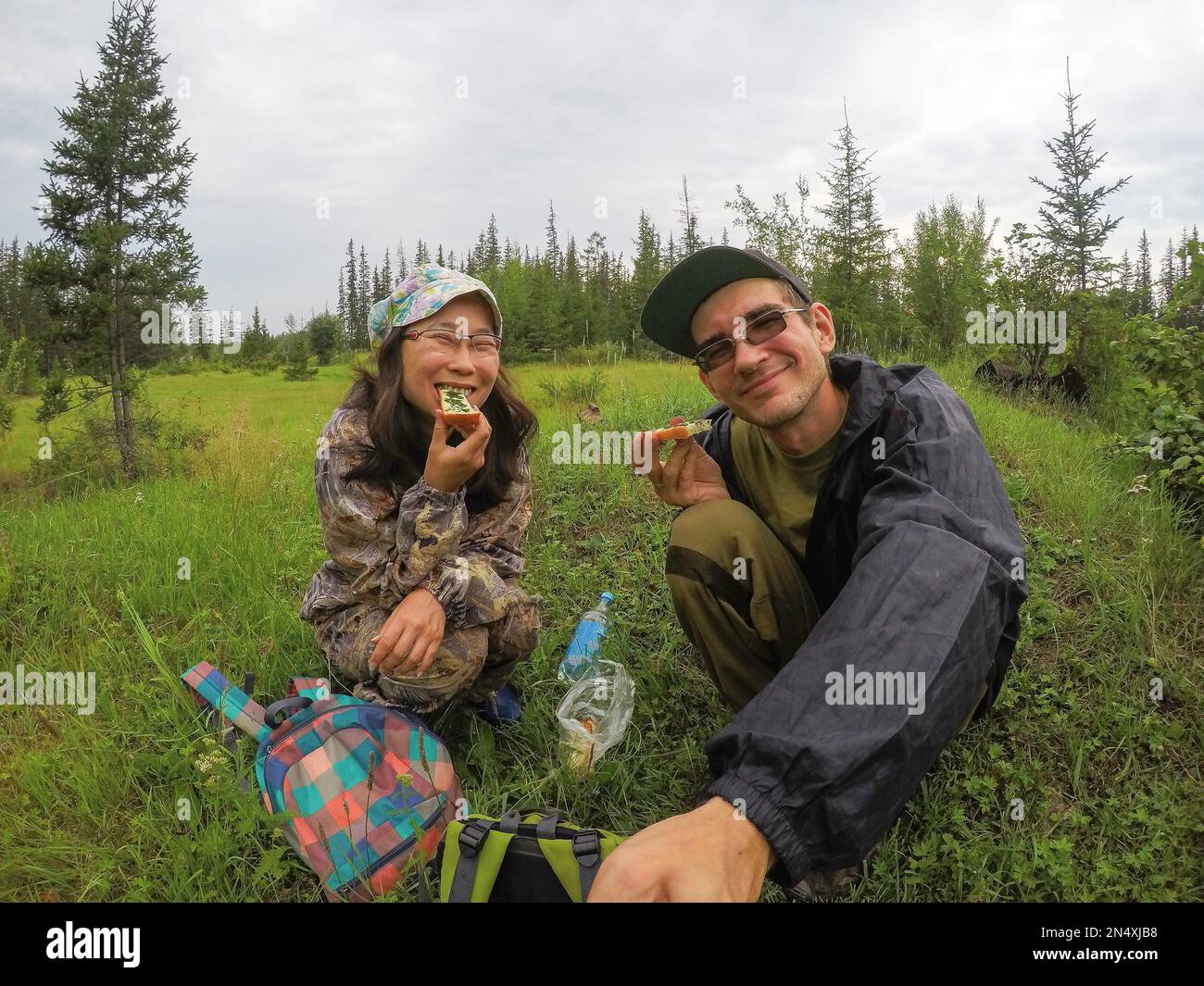Russian man with glasses making a selfie with a happy Asian girl Yakut for a picnic with sandwiches in the Northern taiga wild forest. Stock Photo