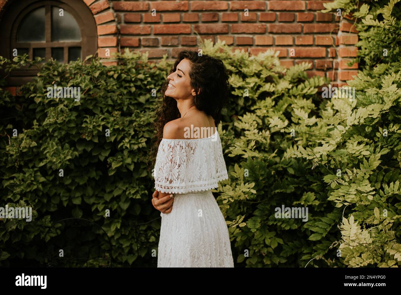 Pretty young woman with curly hair stands in a beautiful garden, dressed in a white flowing dress. She looks relaxed and content Stock Photo