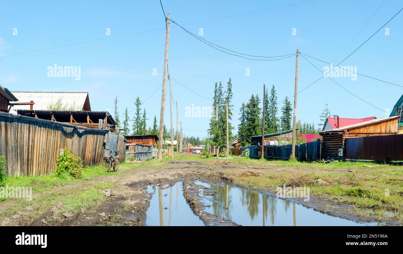 Bright day in the village of ulus Suntar in Yakutia on a residential street with a large puddle and a car in the distance among the wooden houses. Stock Photo