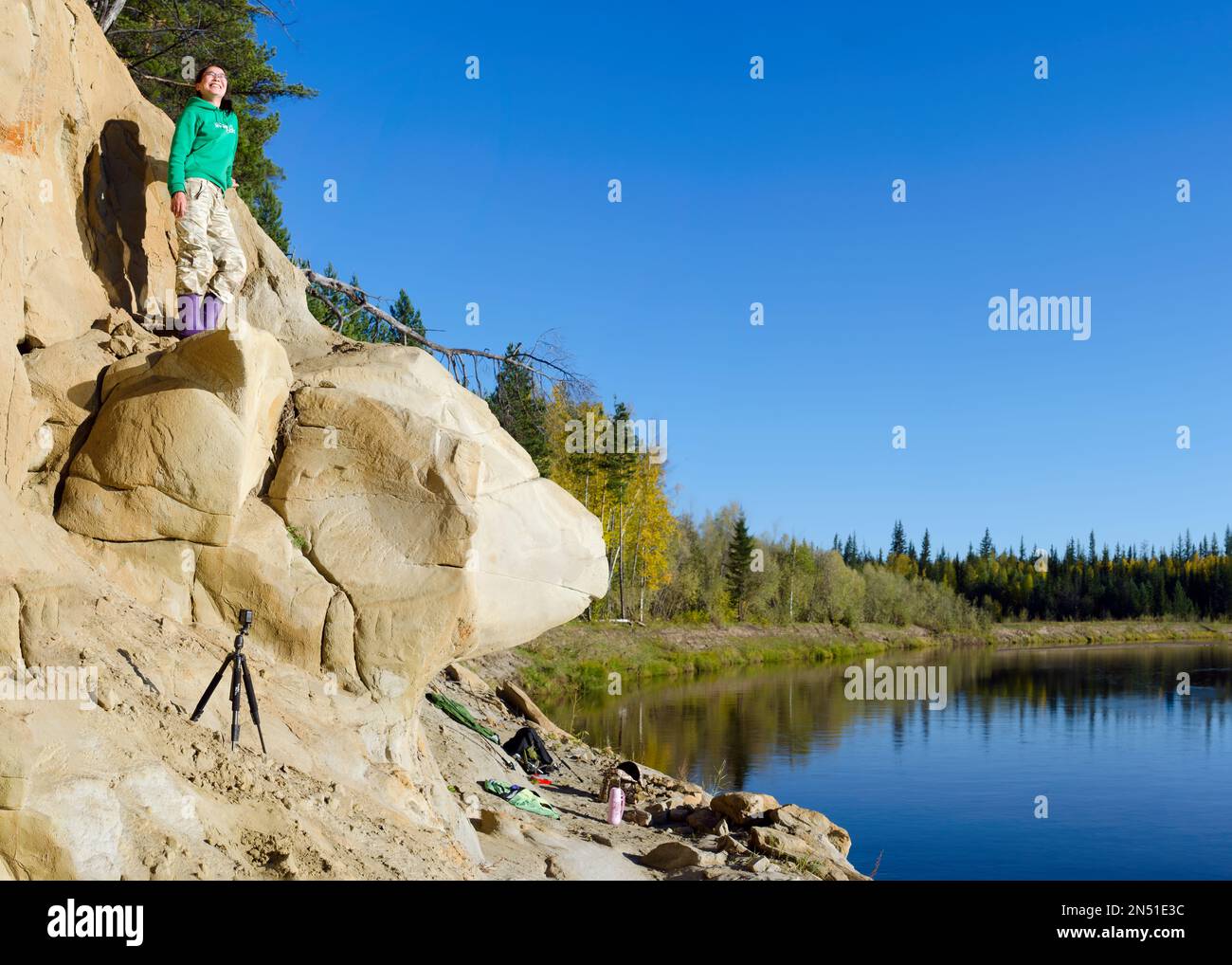 Yakut smiling girl model tourist stands on a clay ledge in the taiga of Northern Yakutia under the trees by the river next to the action camera Stock Photo