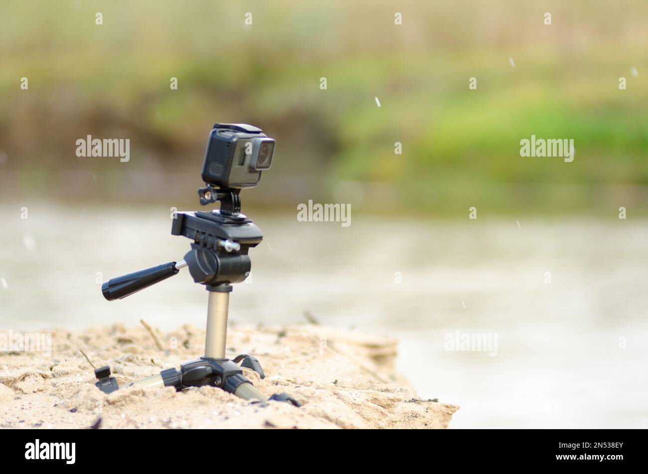 Action camera stands on a small tripod, recording video, on the sandy Bank of the river with grass and rain. Stock Photo