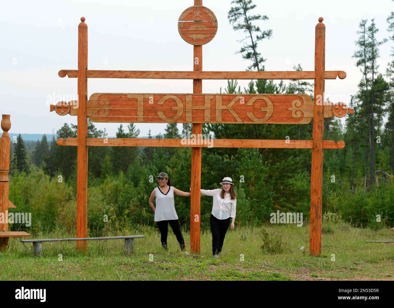 Two friends of an Asian Yakut stand under a wooden sign with the name of the village 'Tenke' in Northern Russia in the taiga forest of Yakutia. Stock Photo