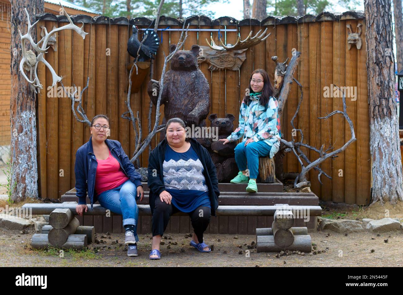 Three colorful girls Asian Yakut sitting on a bench in the zoo at the wooden sculptures of bears and other animals in the village of Ustye in Yakutia. Stock Photo