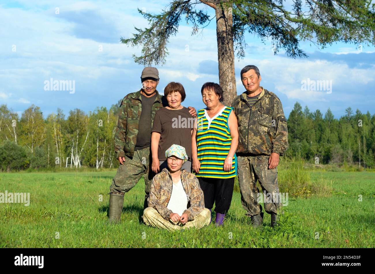 Two Yakut Asian elderly couples men and women with a young girl sitting on the grass pose for a family photo in a field near a tree. Stock Photo