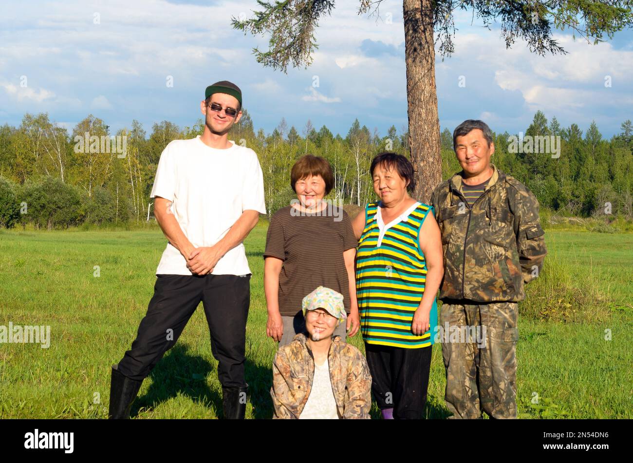 A tall Russian man in boots stands next to a short Asian Yakuts and a girl sitting on the grass posing for a photo in a field in the wild North forest Stock Photo