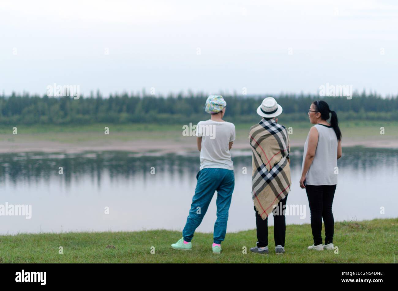 Three girls friends Yakuts, one wrapped in a blanket, stand on the edge of the shore looking at the wild Northern river vilyuj at sunset in the evenin Stock Photo