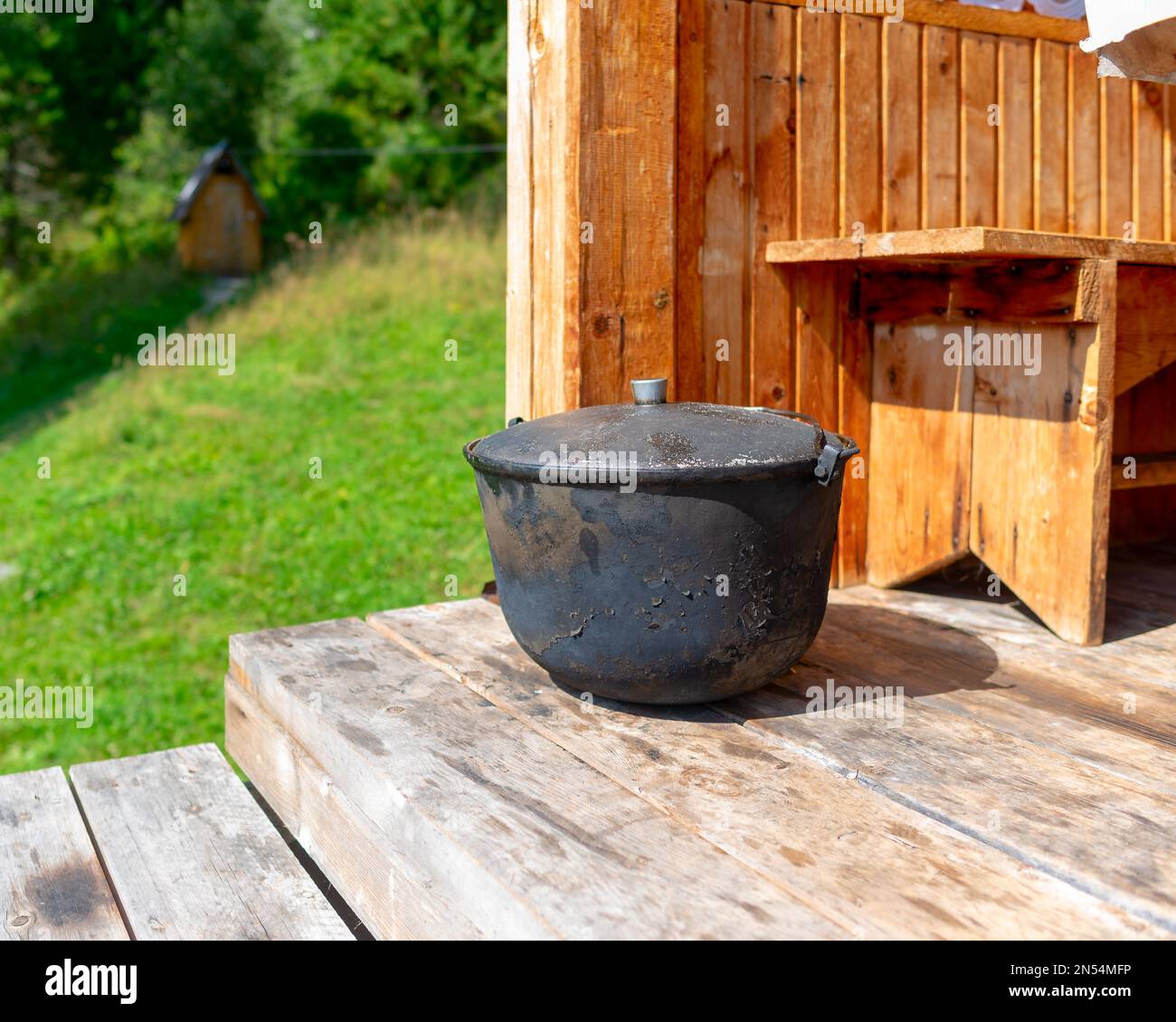 A large, black from soot, the cauldron, the pot stands on the edge of a wooden house a bright day amid toilet and herbs. Stock Photo