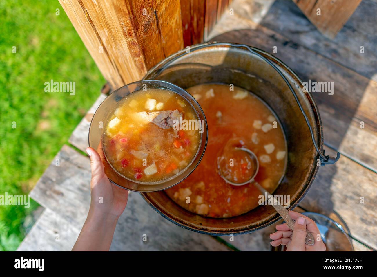 Hands girls cook outdoors lays out soup, lunch with meat and vegetables and potatoes, borscht from a large pot on the wooden floor, in the house next Stock Photo