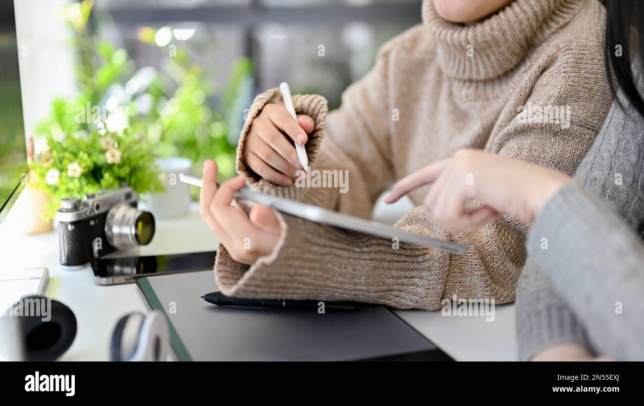 Creative Asian female graphic designer in cozy sweater using digital tablet and discussing work with her colleague in the office. cropped shot Stock Photo