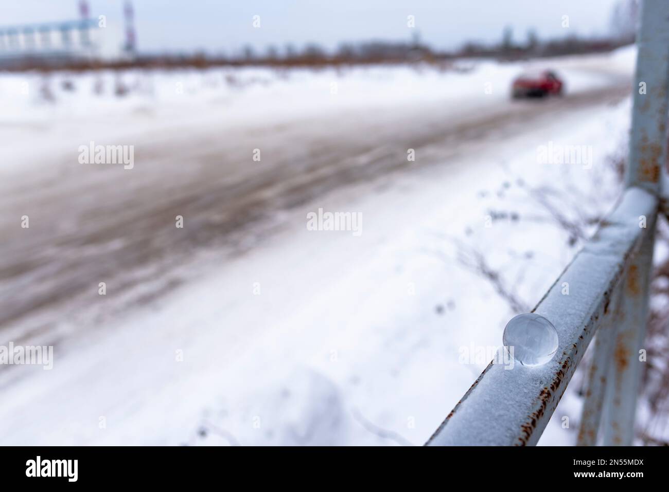 A small glass ball lies in winter on the railing in the snow on the background of a dirty road with a car leaving. Stock Photo