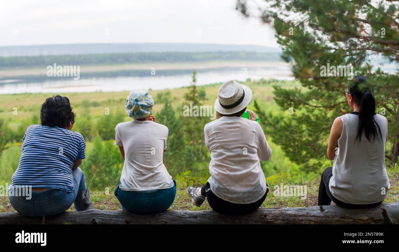 Four girls Yakut friends of tourists sitting resting backs on a log on the mountain overlooking the North river vilyu and taiga. Stock Photo