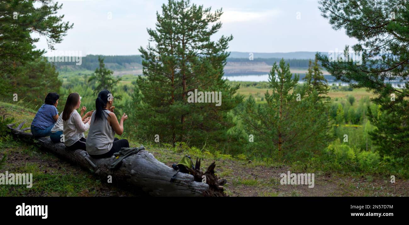 Four Yakut girls friends of tourists sit chatting and gesticulating resting on a log on a mountain overlooking the North vilyu river and the taiga at Stock Photo