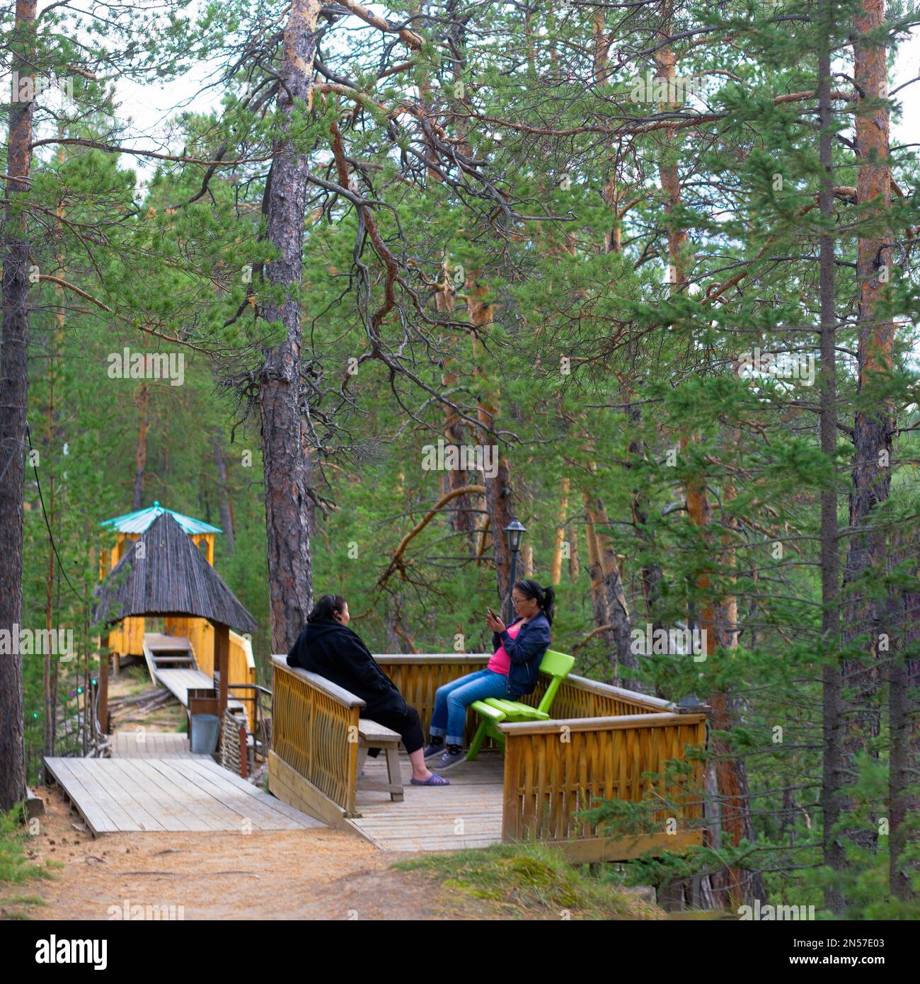 Two Yakut girls are resting on a bench in the forest in the Park playing phone in the Northern taiga. Stock Photo