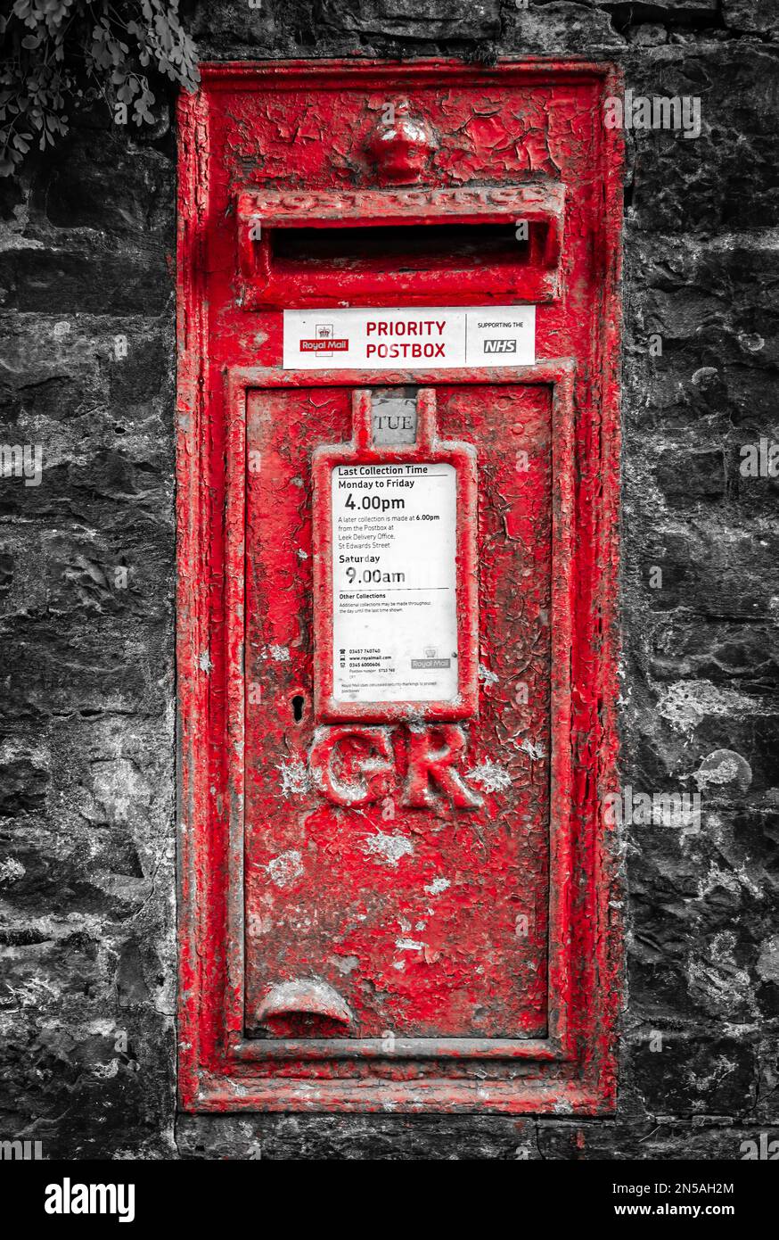 An old bright red wall-set postbox against a black and white background. The postbox dates from King George V, so peeling paint shows its age Stock Photo
