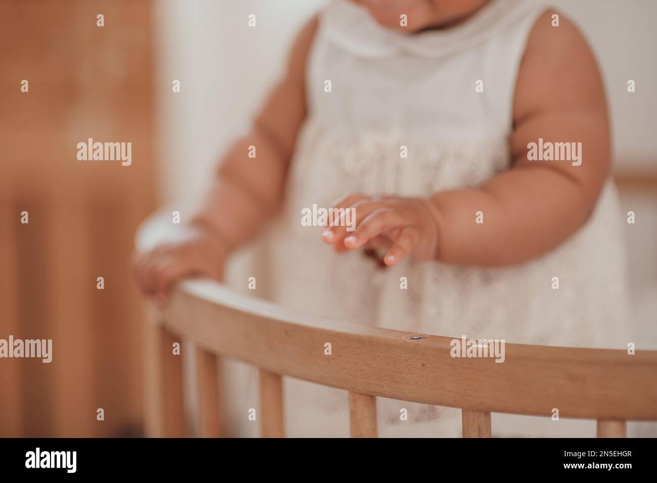Candid authentic baby hands of cute african american diverse female child at home interior of earth colors. Swarthy infant girly stands in kids bed in Stock Photo