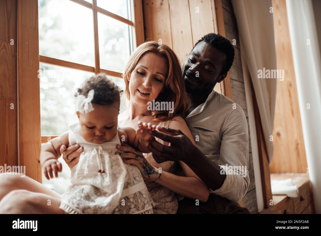 Mixed race family with dark skinned infant daughter spend time together use light window at snug apartment. African american man his fair skin wife an Stock Photo