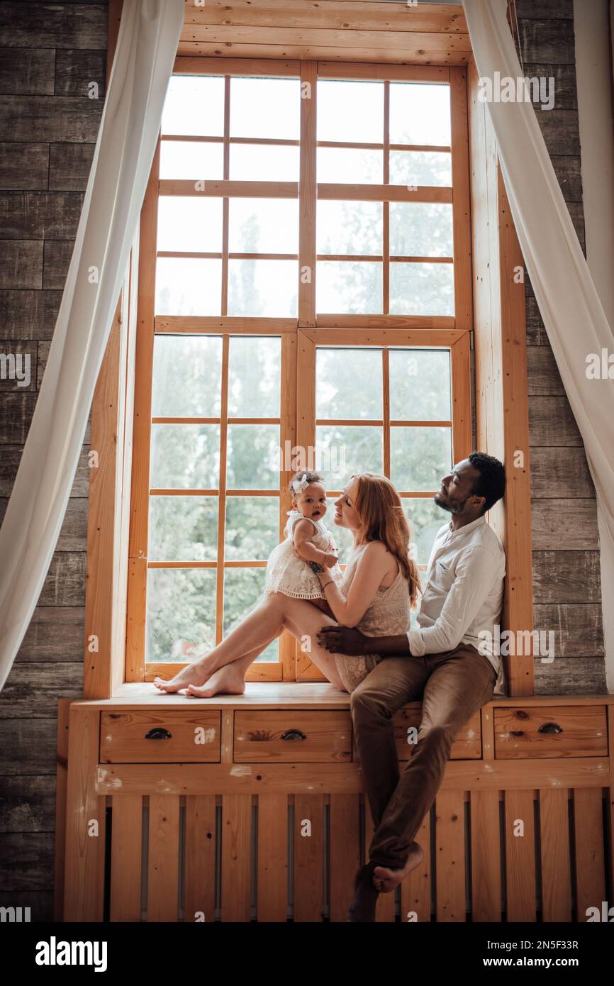 Mixed race family with dark skinned infant daughter spend time together use light window at snug apartment. African american man his fair skin wife an Stock Photo
