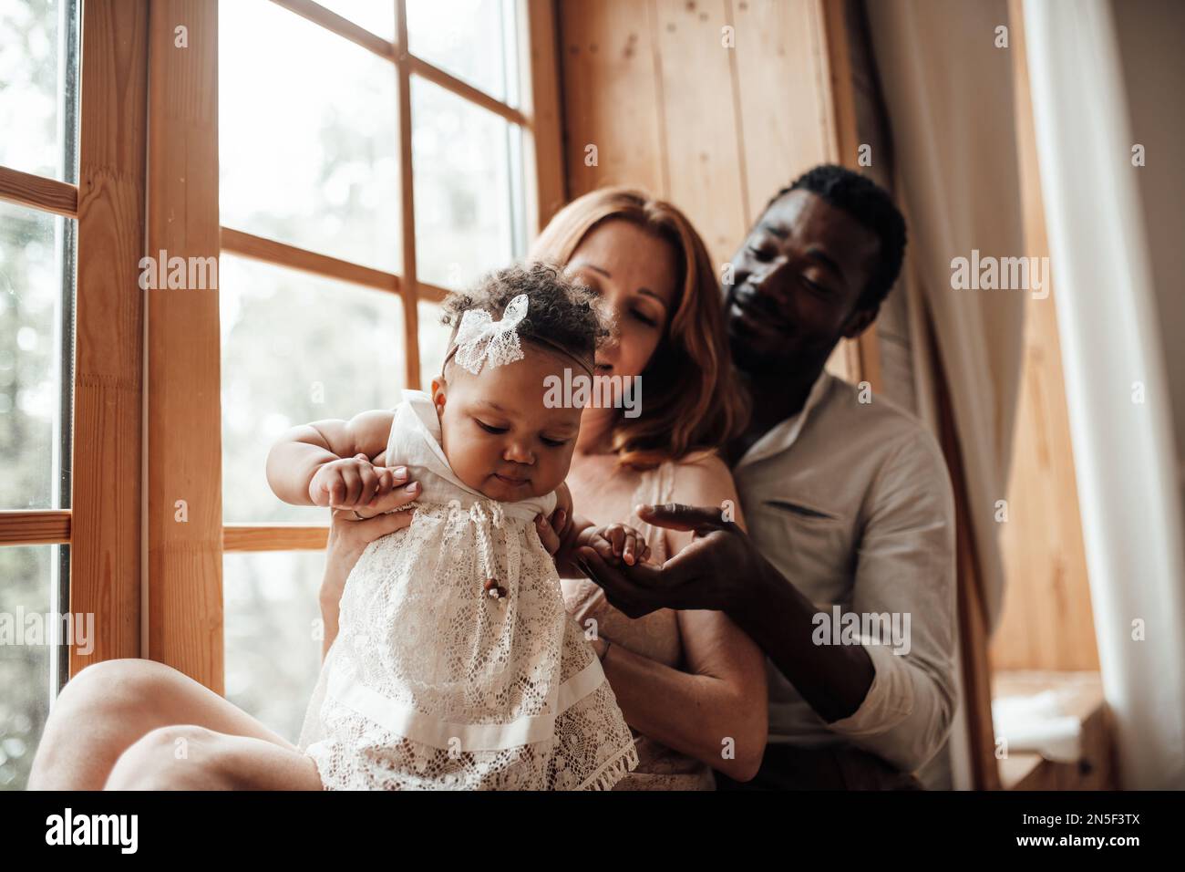 Mixed race family with dark skinned infant daughter spend time together use light window at snug apartment. African american man his fair skin wife an Stock Photo