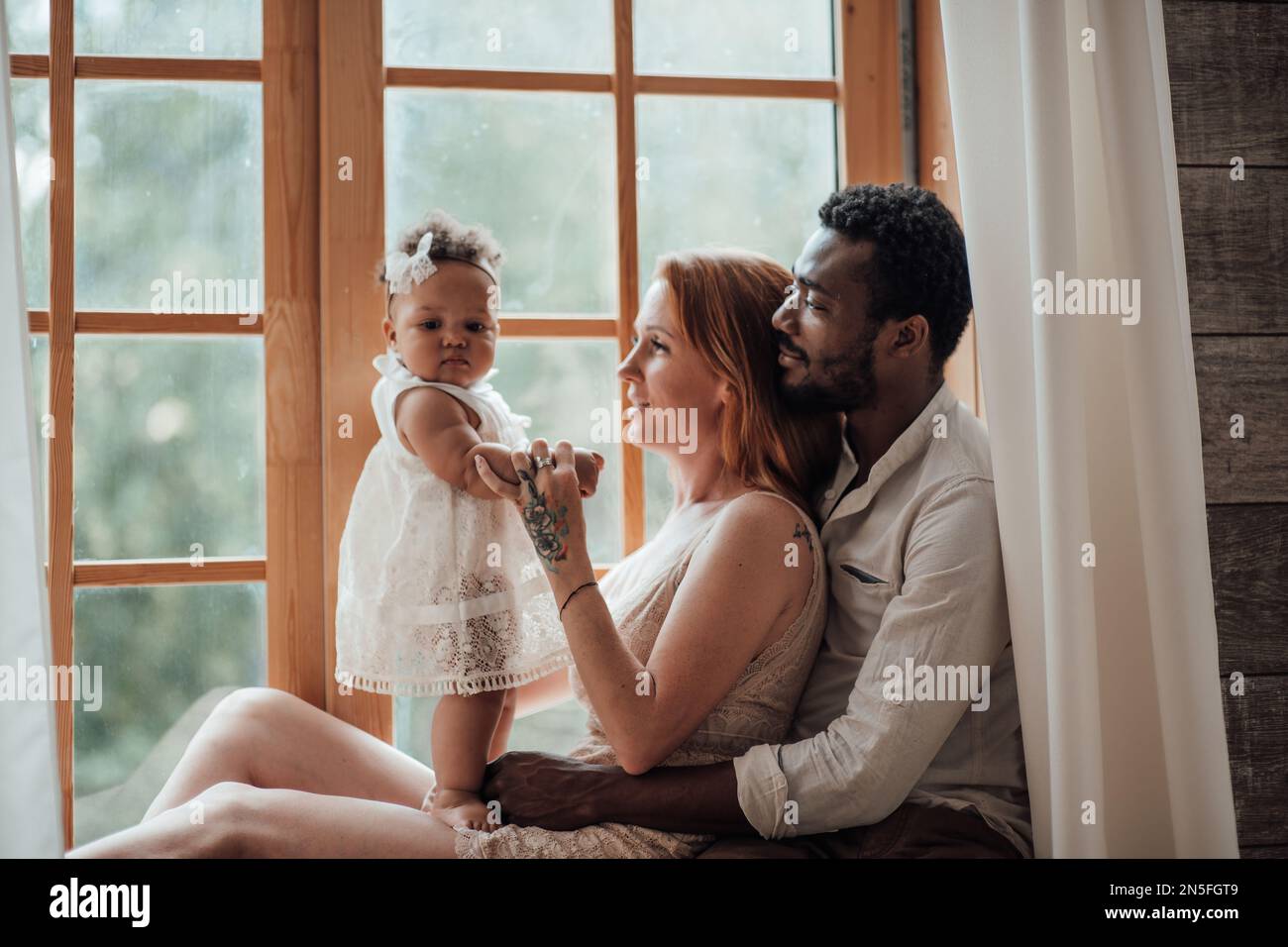 Mixed race family with dark skinned infant daughter spend time together use light window at snug apartment. African american man his fair skin wife an Stock Photo