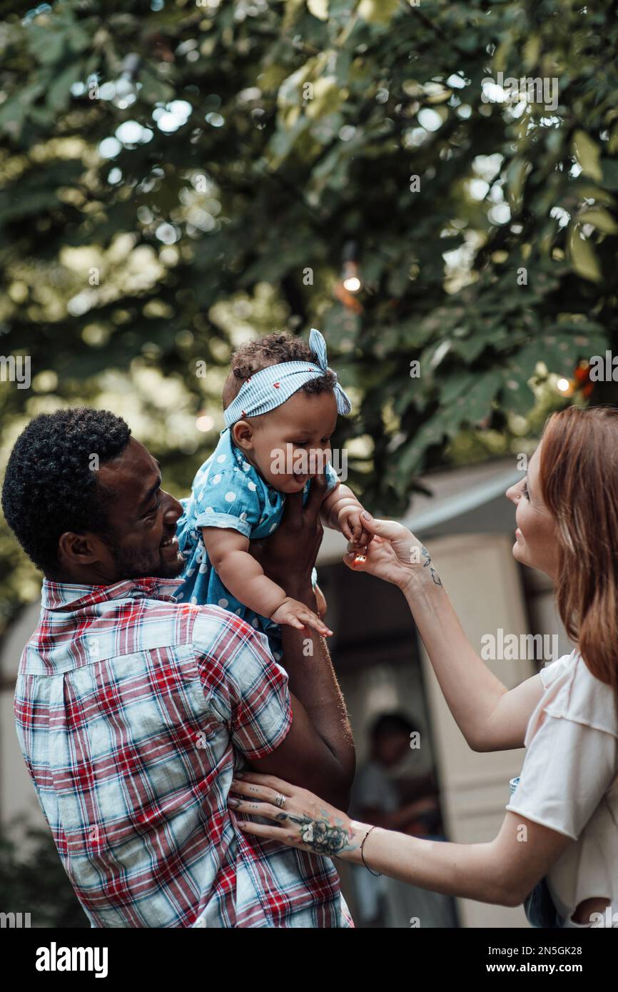 Mixed race family hold dark skinned infant daughter in air spend time together near travel trainer. African american man his fair skin wife and little Stock Photo
