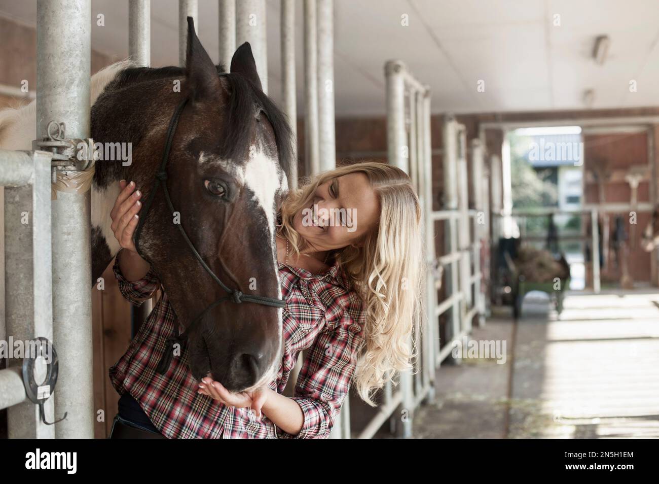 Mid adult woman stroking her horse in barn and smiling, Bavaria, Germany Stock Photo
