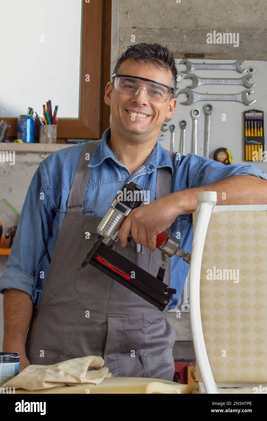 Smiling upholsterer in his workshop with various tools and nail gun after restoring an old chair. Stock Photo