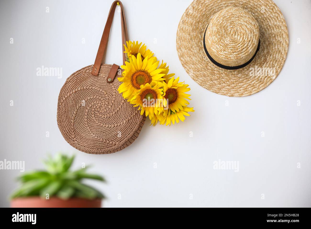 https://c8.alamy.com/comp/2N5HB28/knitted-bag-with-bouquet-of-beautiful-sunflowers-and-straw-hat-on-white-wall-2N5HB28.jpg