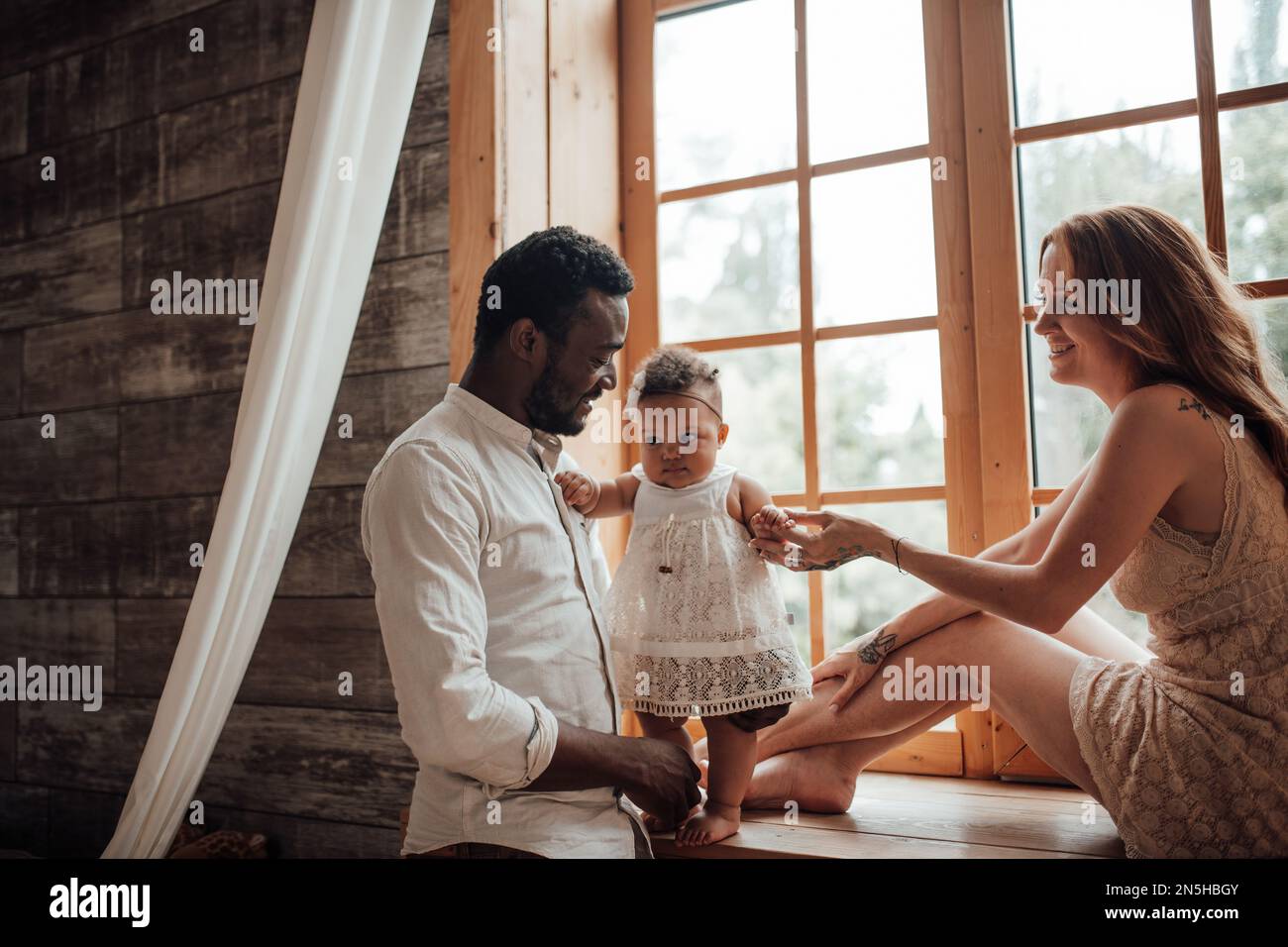 Mixed race family with dark skinned infant daughter spend time together use light window at snug apartment. African american man his fair skin wife an Stock Photo