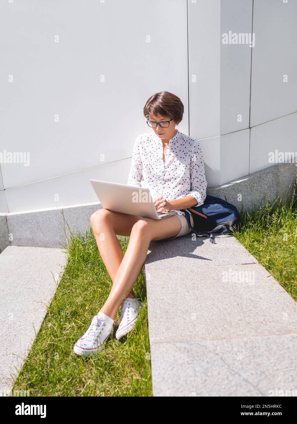 Woman sits with laptop on urban park bench. Freelancer at work. Student learns remotely from outdoors. Modern lifestyle. Summer vibes. Stock Photo