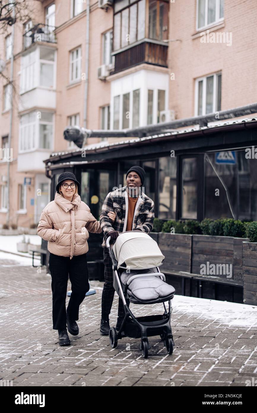 Happy interracial family walk on street and push baby carriage. Concept of interracial family and unity between different human races. Stock Photo