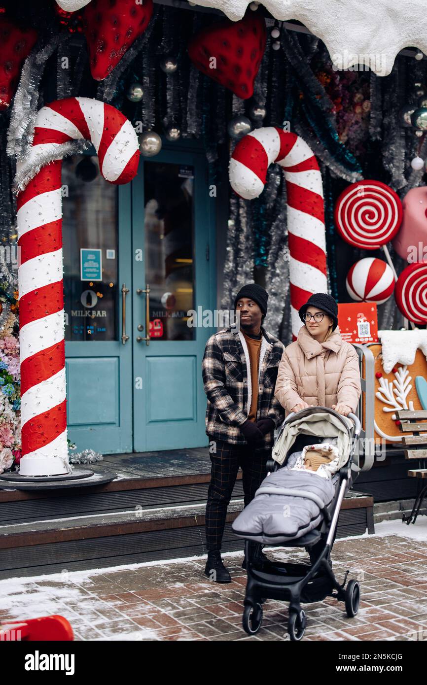 Happy interracial family walks at Christmas fair with baby carriage. Concept of interracial family and unity between different human races. Stock Photo