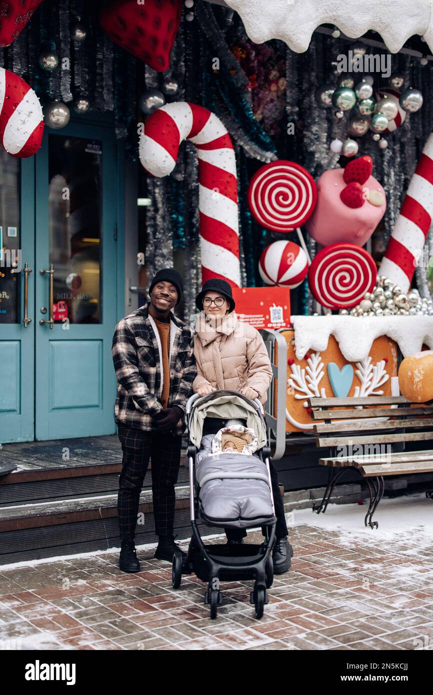 Happy interracial family walks at Christmas fair with baby carriage. Concept of interracial family and unity between different human races. Stock Photo