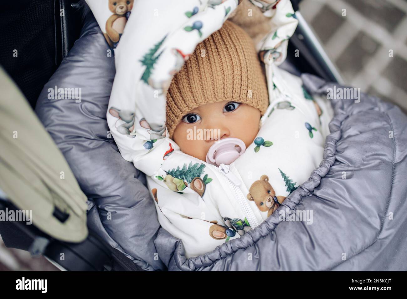 Dressed and wrapped infant from interracial marriage lies down in baby carriage during walk in winter. Concept of unity between different human races. Stock Photo