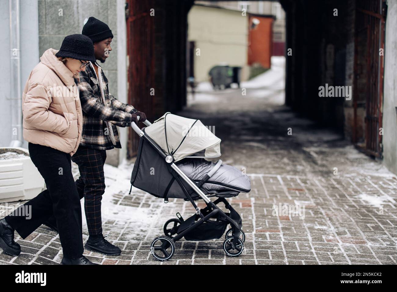 Happy interracial family walk on street and push baby carriage. Concept of interracial family and unity between different human races. Stock Photo