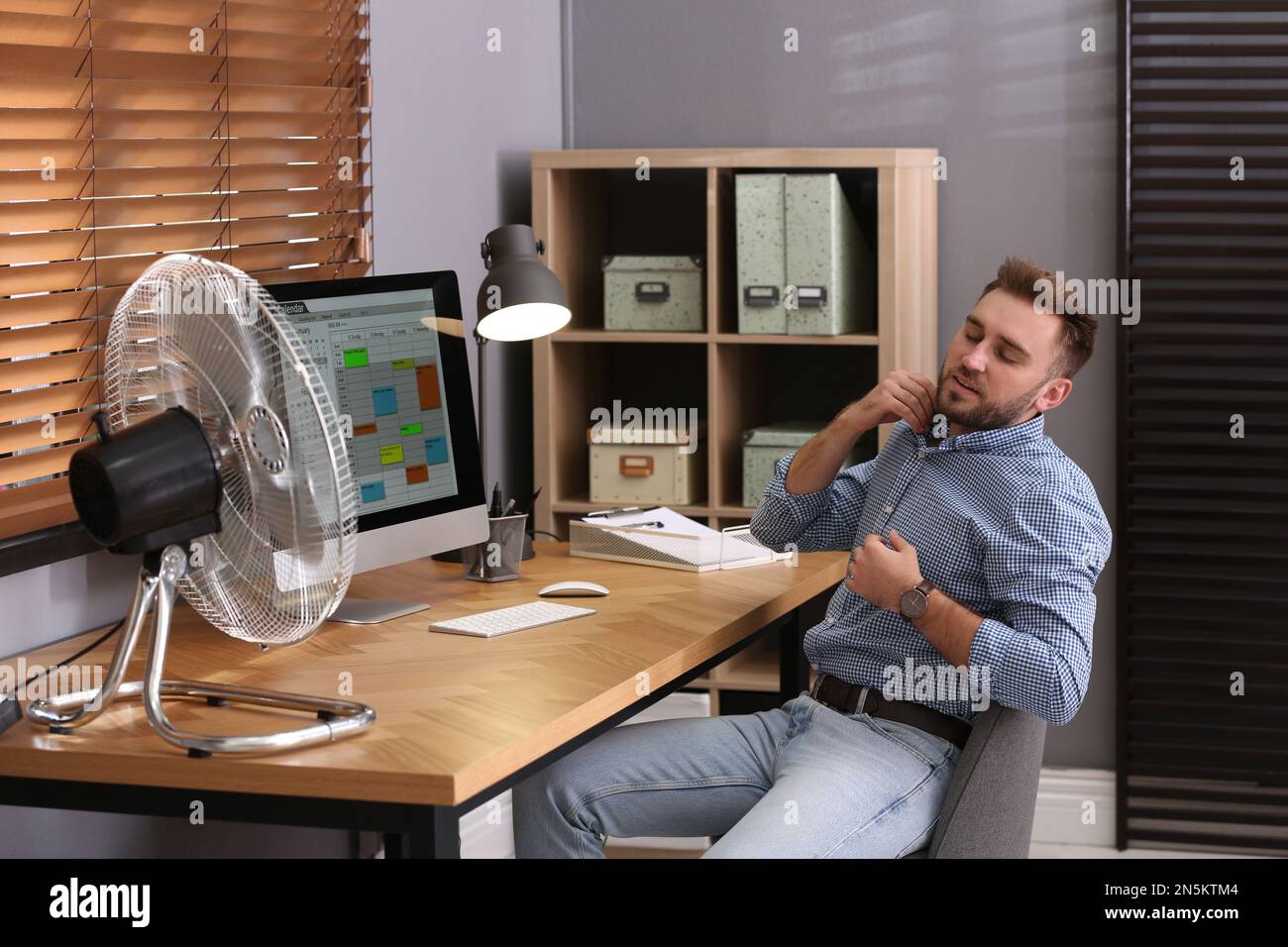 Man suffering from heat in front of fan at workplace Stock Photo