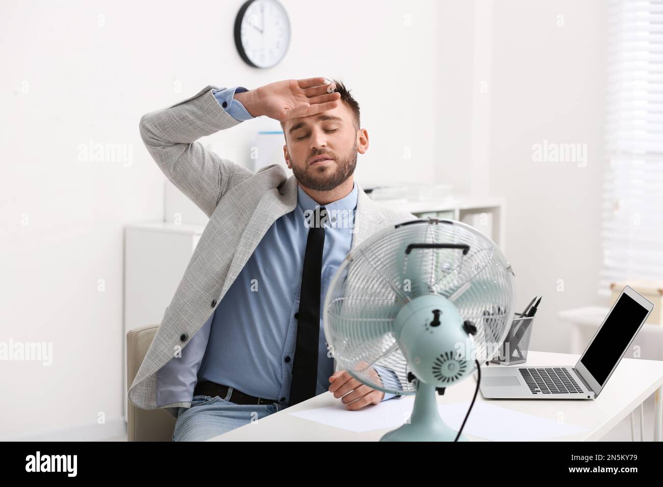 Man suffering from heat in front of fan at workplace Stock Photo