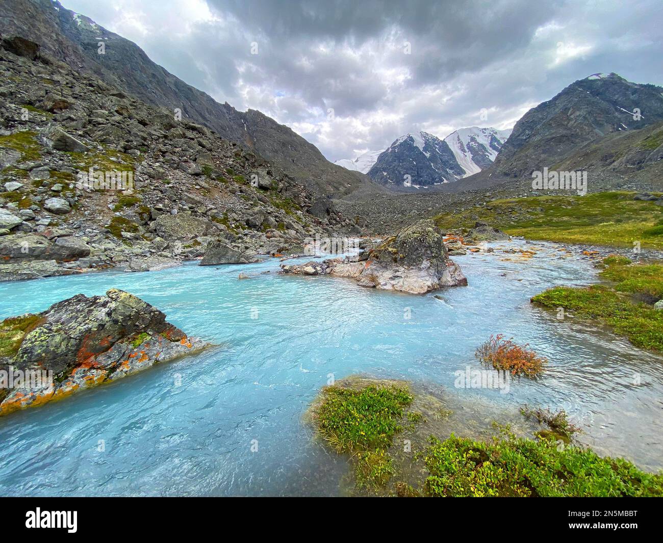 Alpine turquoise stream flows among orange stones from mountains with glaciers in Altai in Siberia. Stock Photo