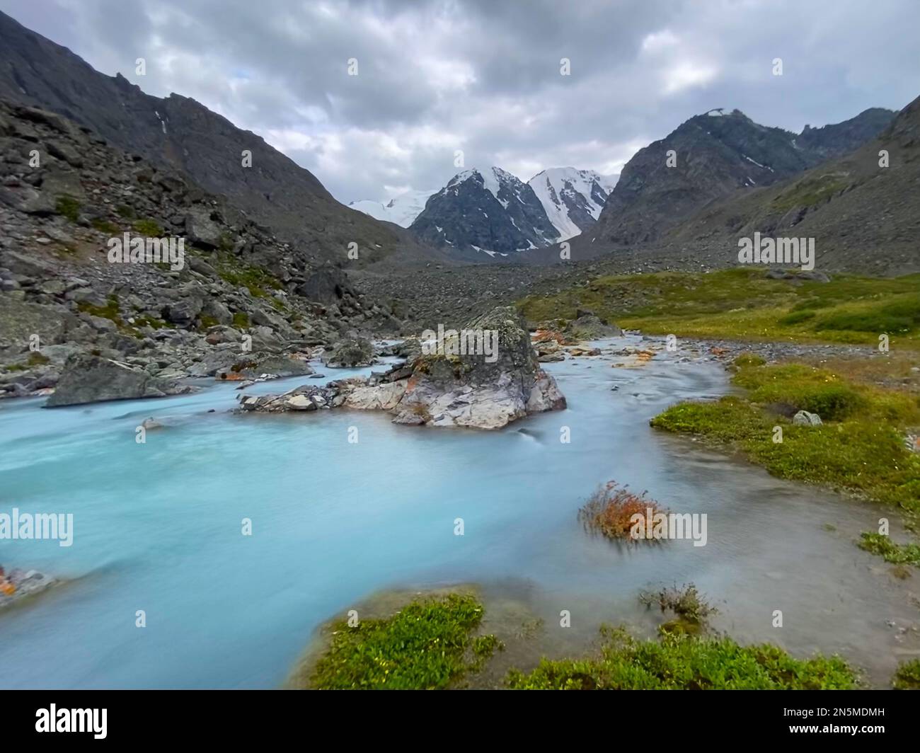 Alpine turquoise stream flows among orange stones from mountains with glaciers in Altai in Siberia during the day. Stock Photo