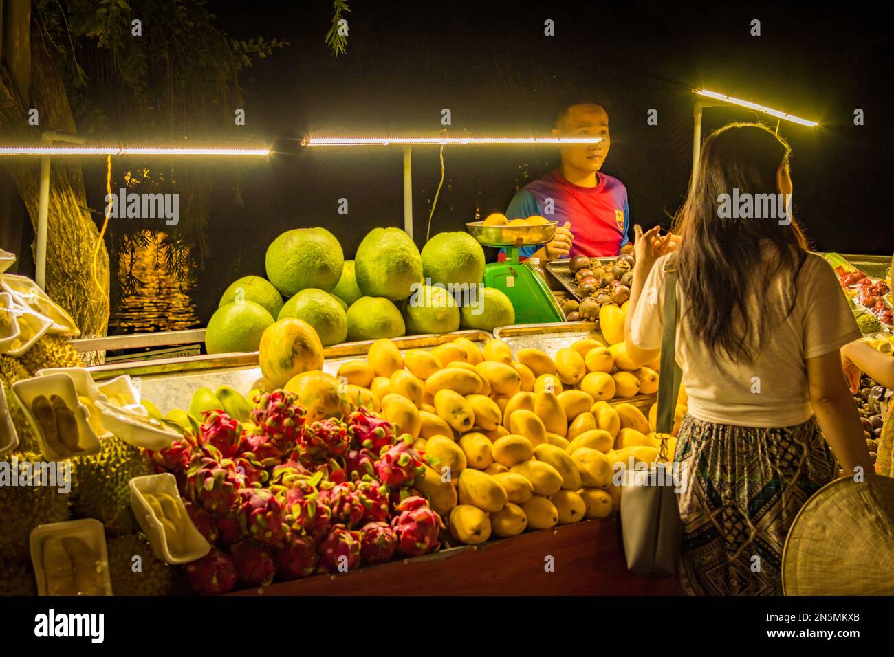 Woman buying food at a fruit and vegetable stall by night in Hoi An, Vietnam. Stock Photo
