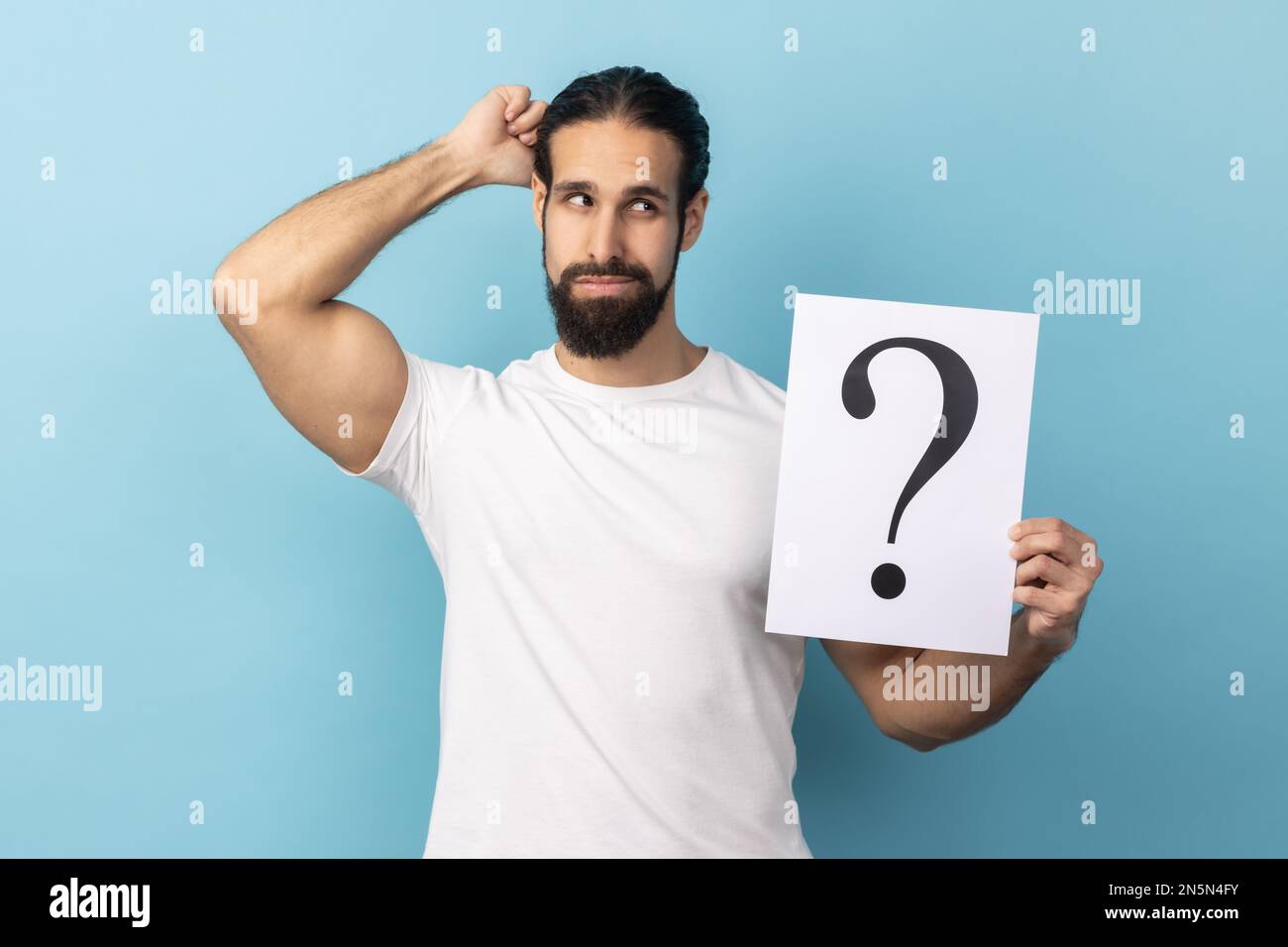 Portrait of puzzled man with beard wearing white T-shirt holding question mark, finding smart solution, asking for advice, looking away. Indoor studio shot isolated on blue background. Stock Photo
