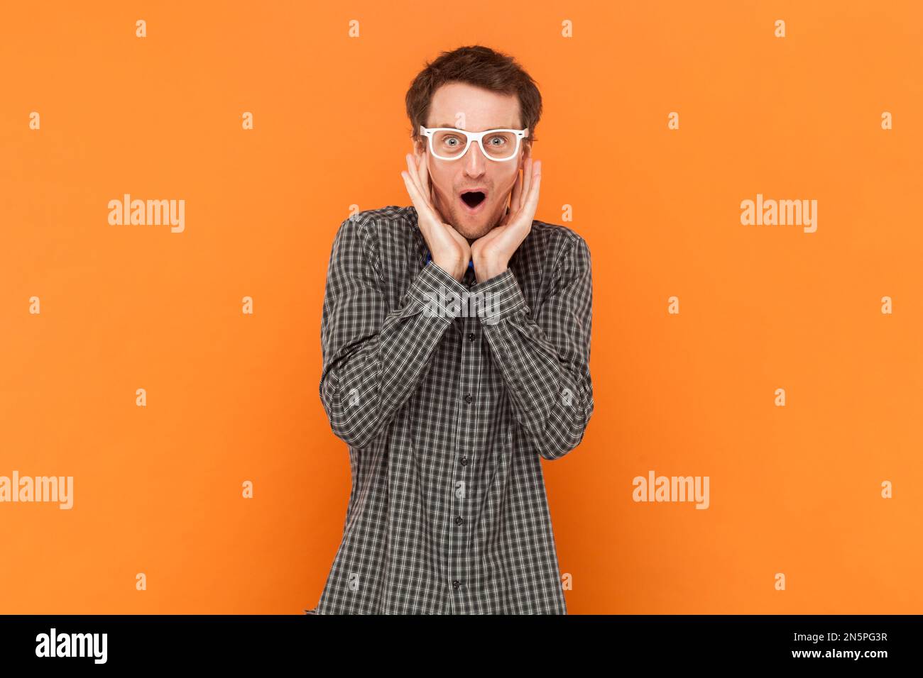 Amazed man nerd doesnt believe his success, keeps hands on head, stares at camera, says omg or wow, wearing shirt with blue bow tie and white glasses. Indoor studio shot isolated on orange background Stock Photo