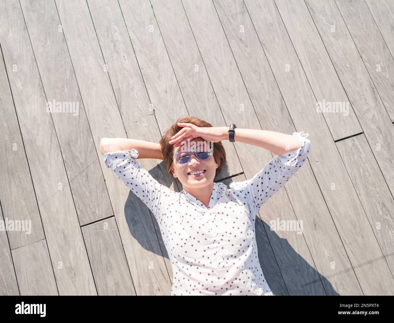 Woman with short hair and freckles poses in colorful sunglasses. Smiling woman is lying on wooden background. Summer vibes. Sincere emotions. Stock Photo