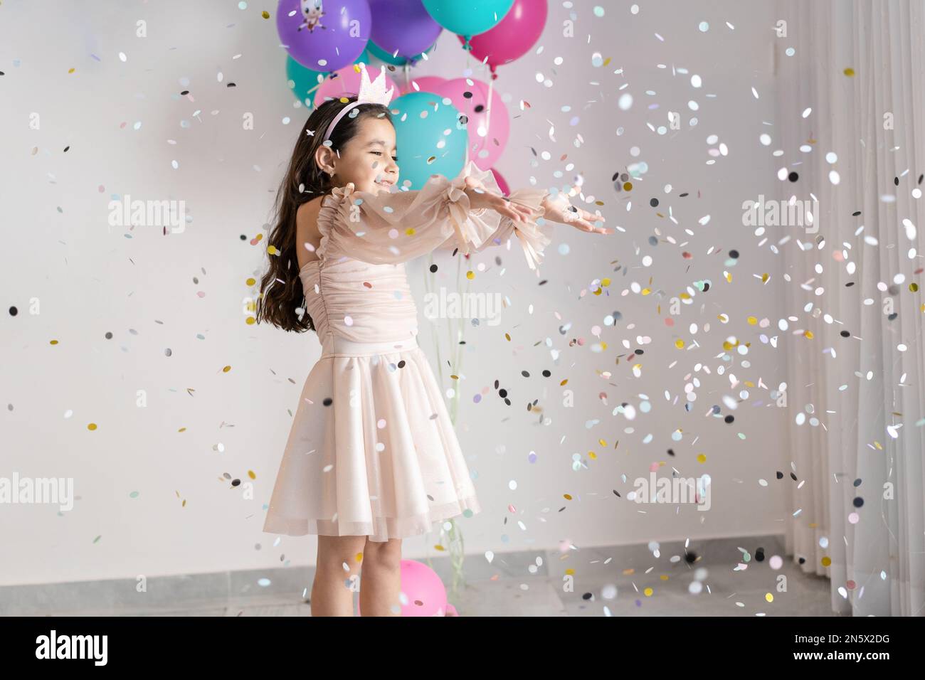 A little brunette girl in a pink dress and crown smiling and throwing colorful eco-friendly paper confetti, confetti falling down on her. White background full of paper circles. Stock Photo