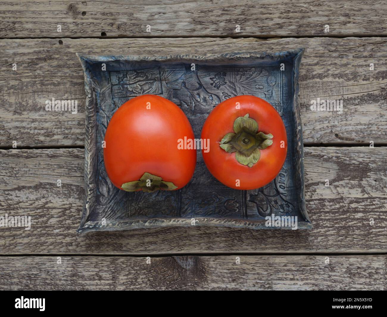 Persimmon fruits on blue ceramic tray on wooden background, close up table top view. Detailed photo of kaki fruit with leaf, photo directly  above. Stock Photo