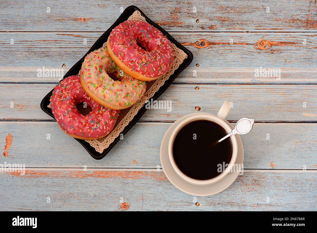 A cup of hot black coffee on a saucer and a plate with three donuts in white and pink glaze on a wooden table. Top view, flat lay. Stock Photo